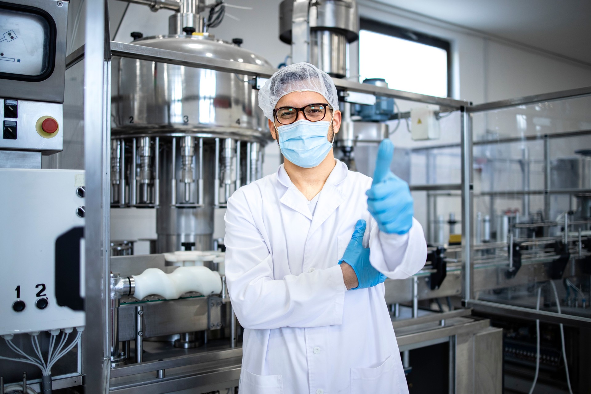 Portrait of technologist or worker in sterile white clothing standing by automated industrial machine in pharmaceutical company or factory.